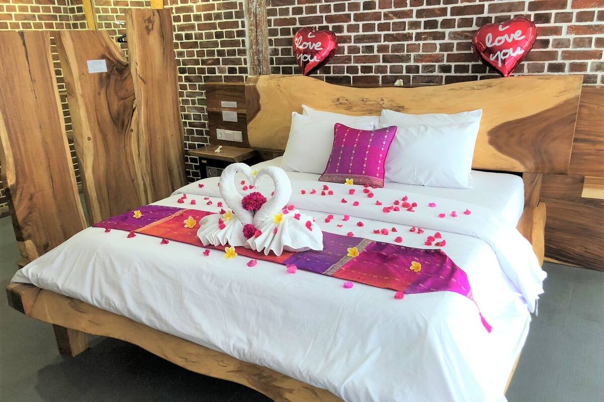 Una cama con cabecera de madera, ropa de cama blanca, una colcha morada y rosa, y una disposición de toallas en forma de corazón.