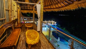 Una terraza interior de madera con bancos y techo de paja, con vista a una fuente de agua.