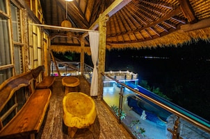 Una terraza interior de madera con bancos y techo de paja, con vista a una fuente de agua.
