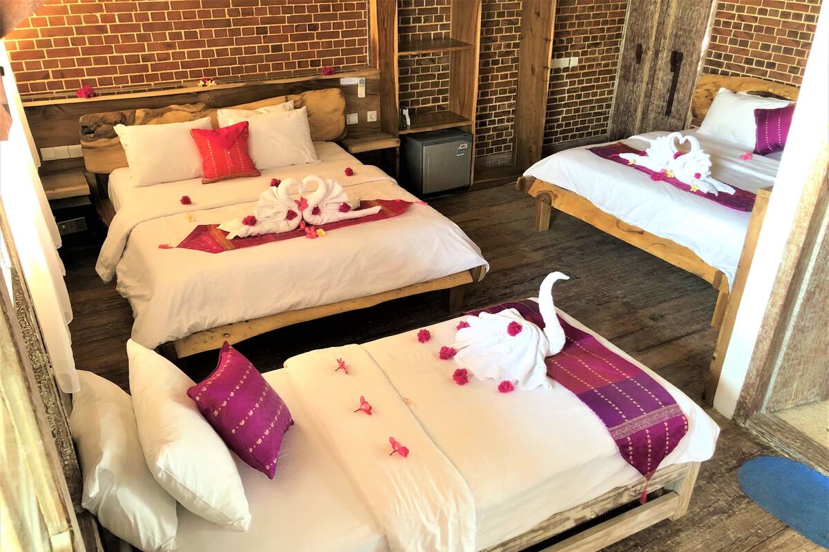 Habitación de hotel con dos camas, cada una con sábanas blancas y almohadas moradas, y una pared de ladrillo.