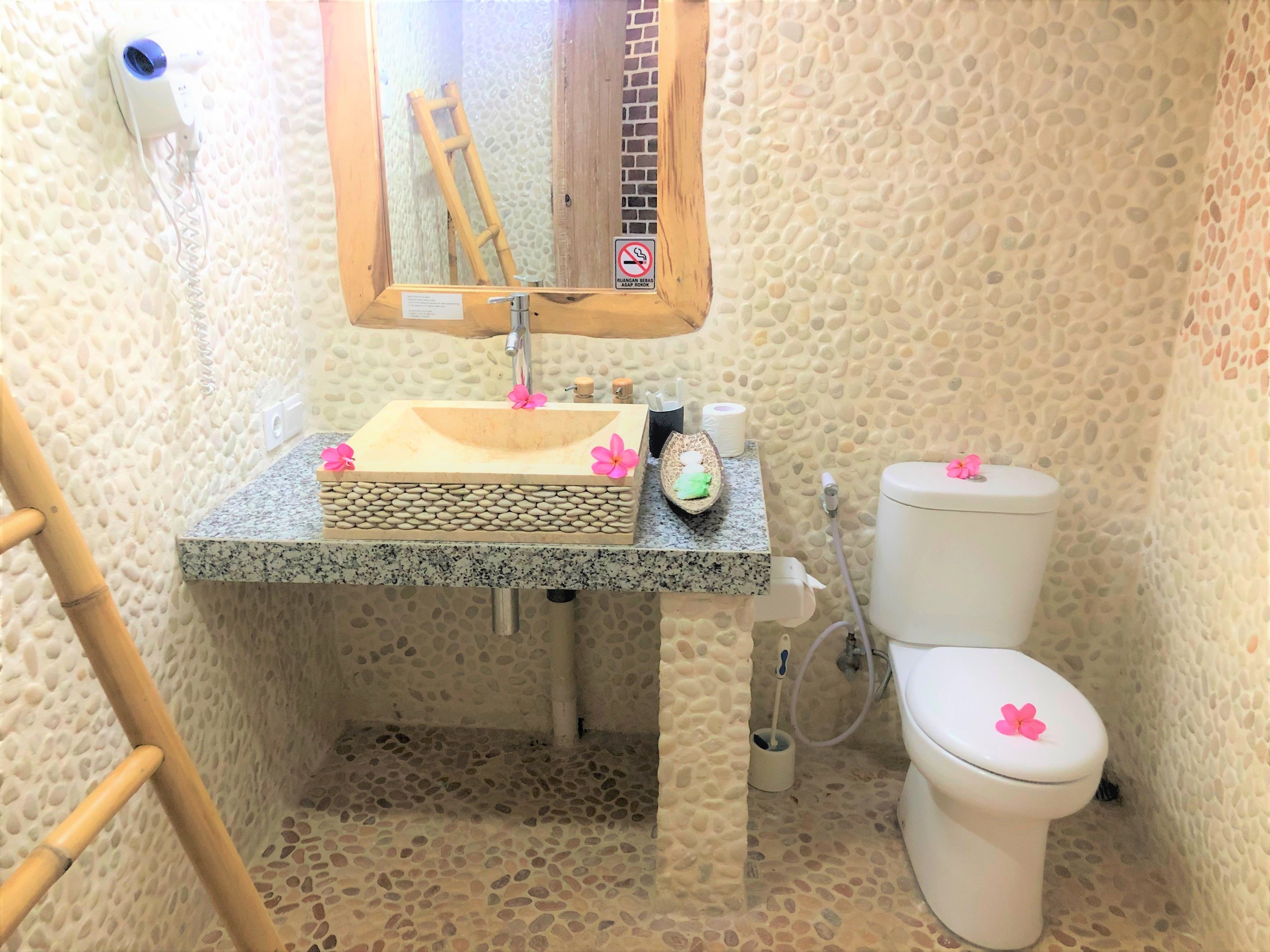A bathroom with a pebble-textured wall, a granite sink, a wooden-framed mirror, a toilet, and a bamboo ladder.