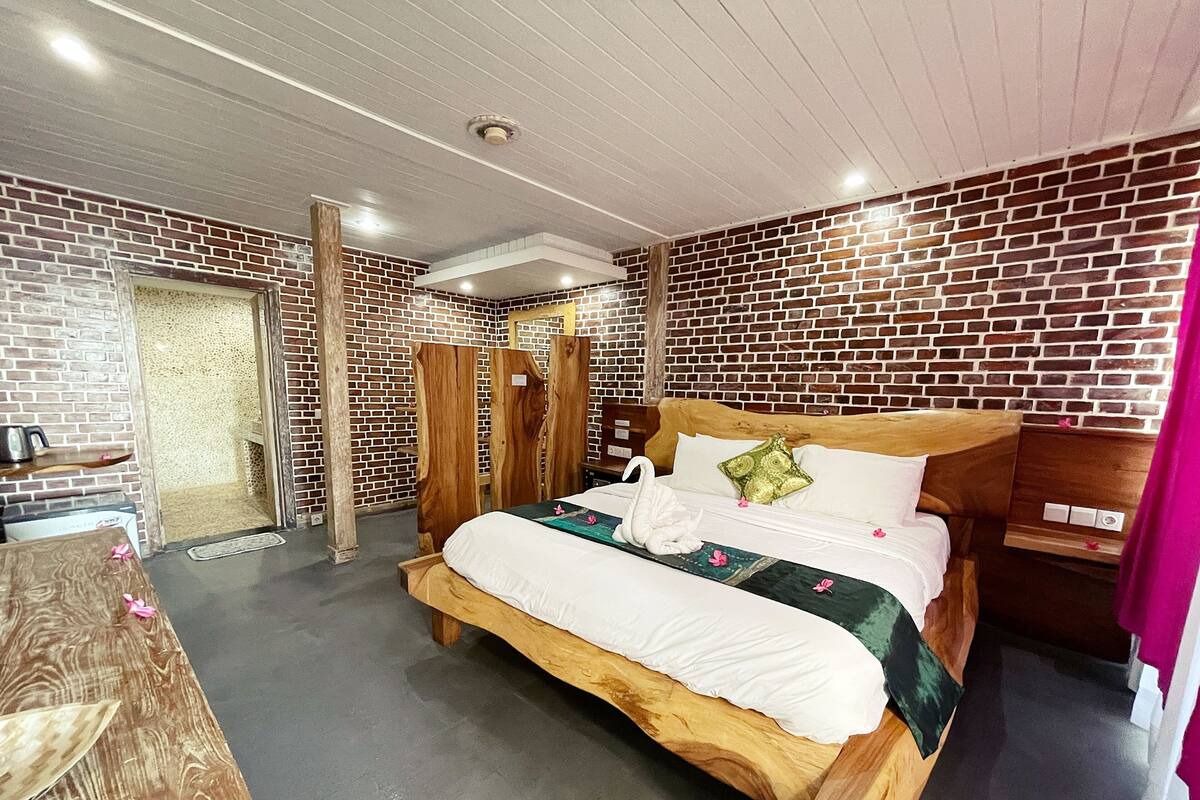 Una habitación con pared de ladrillo, una cama de madera con ropa blanca, una mesita de noche de madera y una pequeña mesa con una silla.