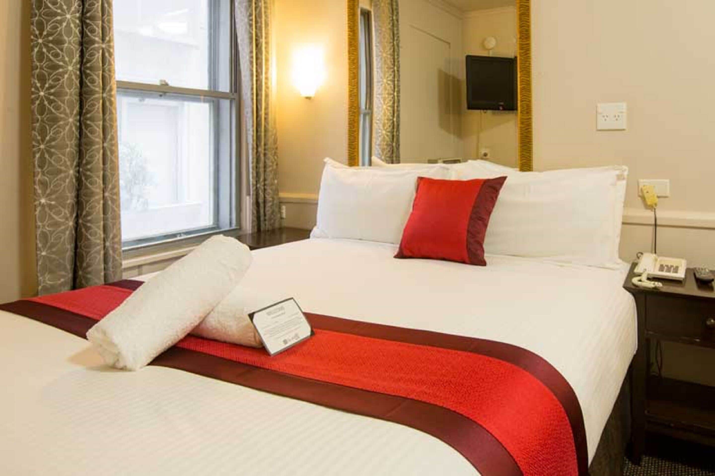 Een hotelkamer met een groot bed, rood-wit beddengoed, een nachtkastje met telefoon en een raam met gordijnen.