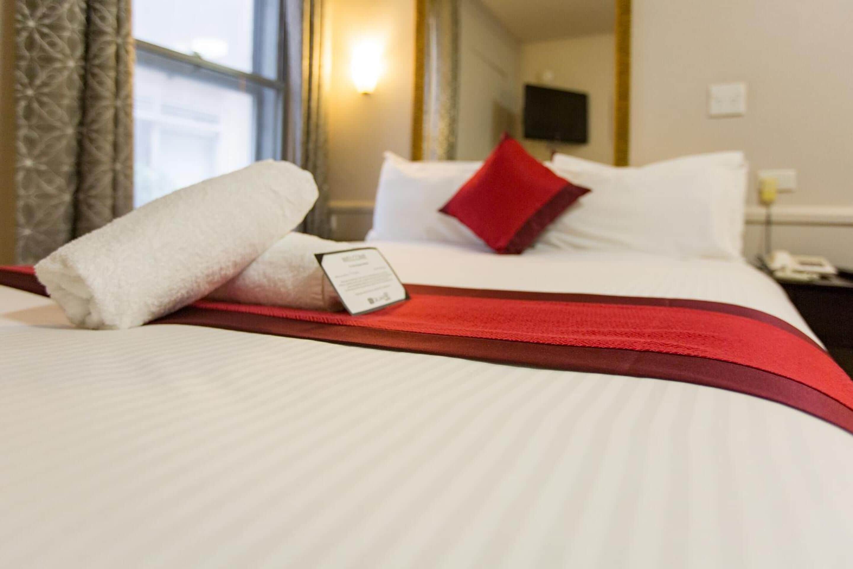 A hotel room with a neatly made bed, a red throw blanket, and a white pillow.