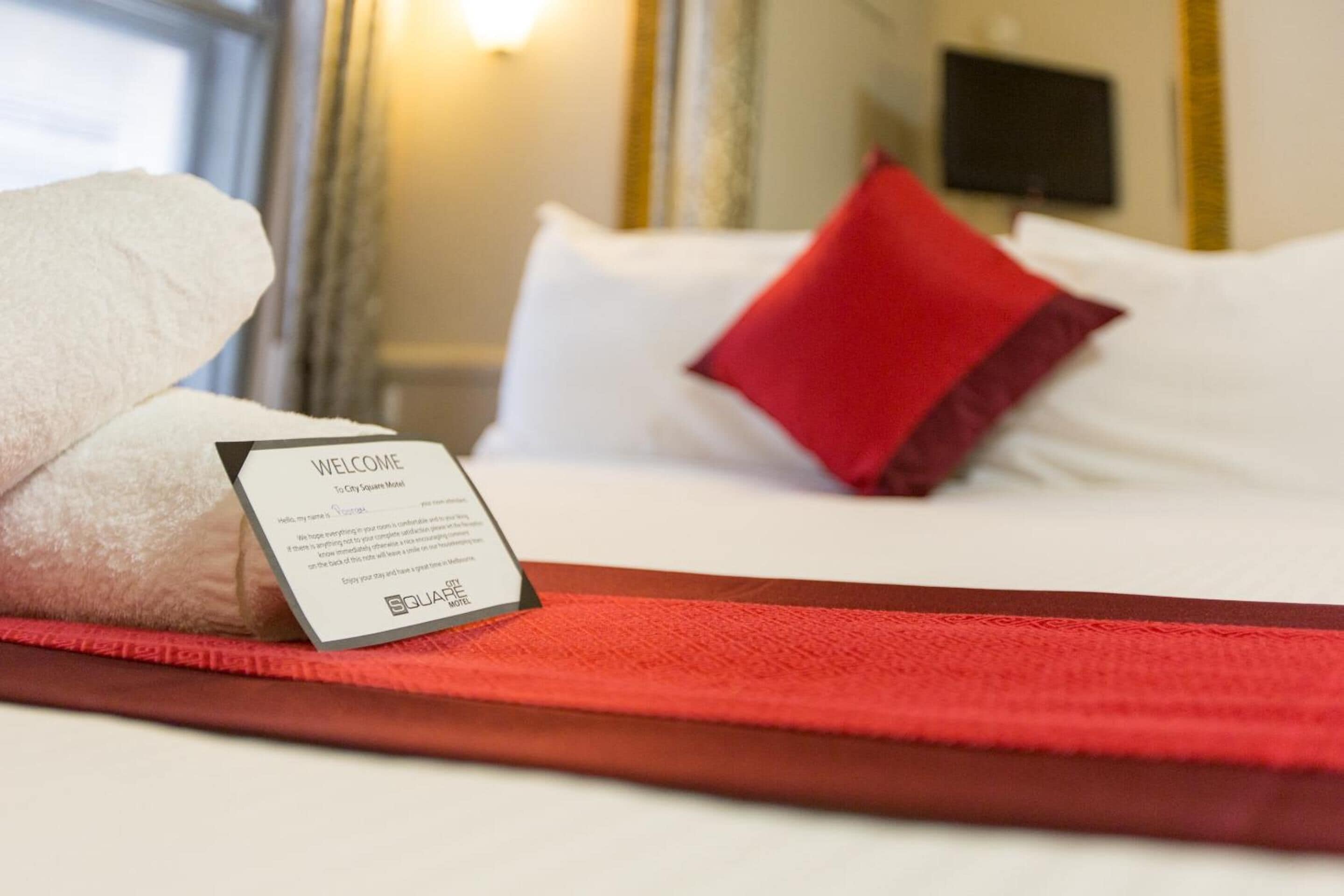 A neatly made bed with a red pillow, a welcome sign, and a red throw blanket.