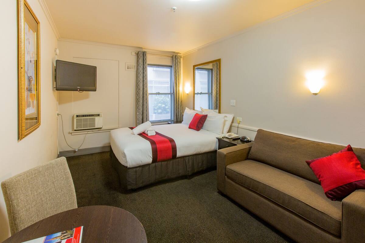 A hotel room with a bed, a sofa, a TV, an air conditioning unit, and a mirror.