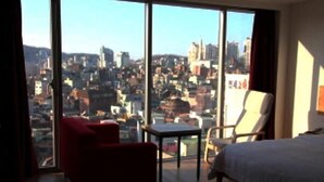 A hotel room with a large window offering a city view, a bed, a red armchair, and a small table. - Lavilla Hotel (Seoul)