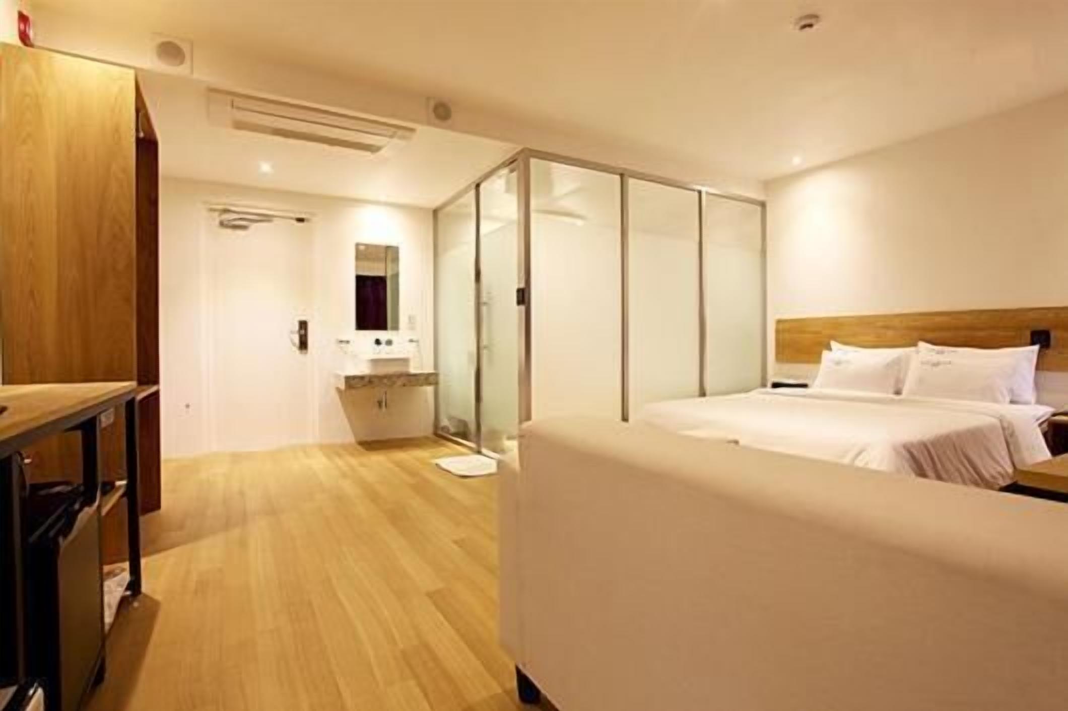 Une chambre d’hôtel avec un plancher en bois, un lit avec du linge de maison blanc, une table de chevet, un miroir et une porte coulissante en verre donnant sur une salle de bain.