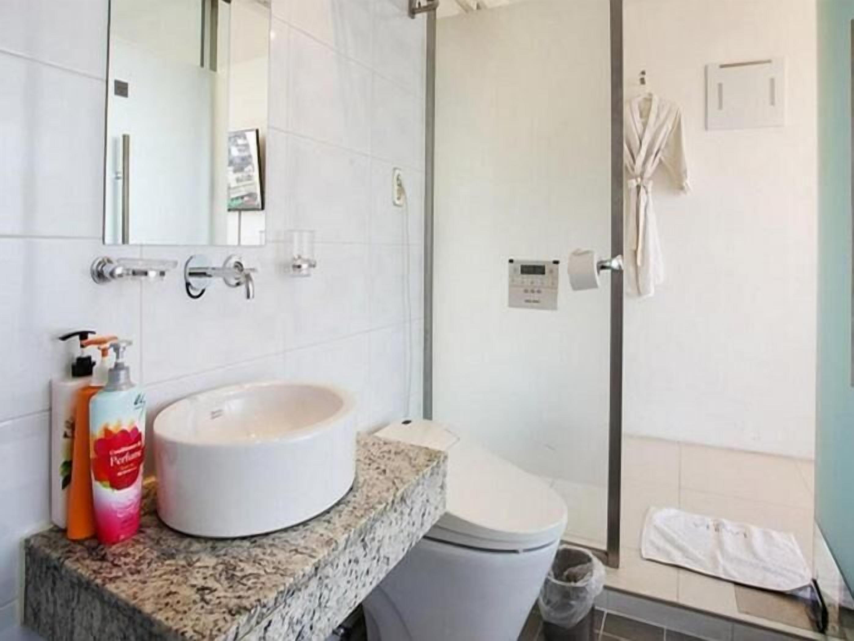 A modern bathroom with a granite countertop, a white sink, and a toilet. There is a shower area with a glass door and a towel hanging on the wall.