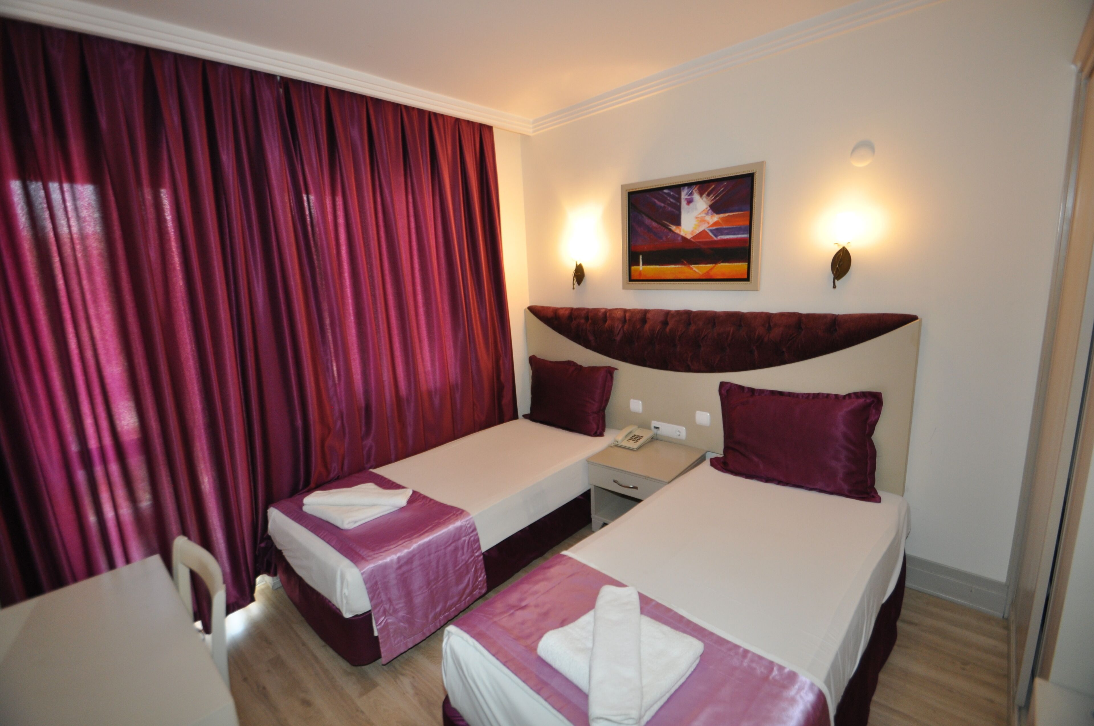 A hotel room with two beds, a desk, a chair, a painting on the wall, and a window with curtains.