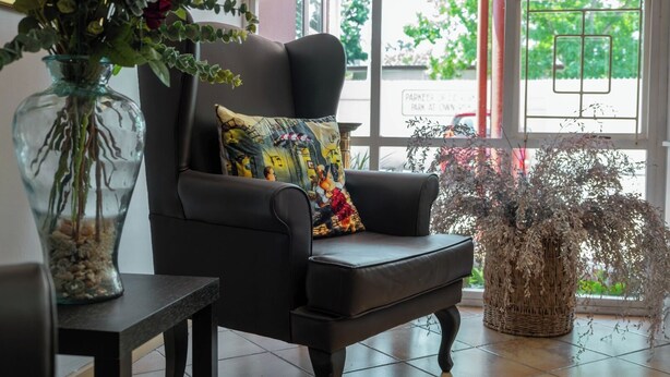A black leather armchair with a colorful pillow, a glass vase with plants, and a wicker basket with dried foliage. - Broadway Guesthouse (Cape Town)