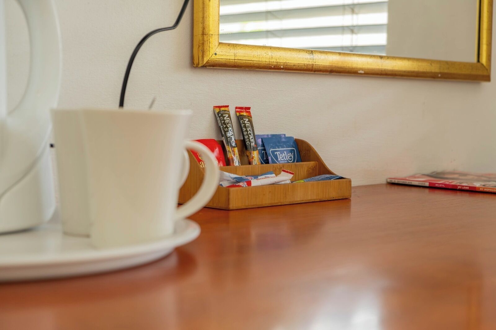 Una taza blanca sobre un plato, una bandeja de madera con varios objetos, y un espejo en un marco colgado en la pared.