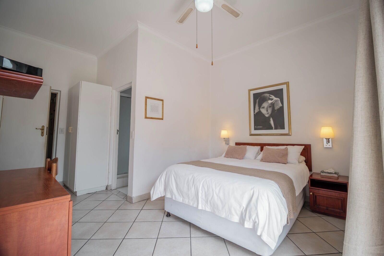 Una habitación de hotel con cama, lámparas de noche, un cuadro enmarcado en la pared, un aparador de madera y una puerta que da a otra habitación.