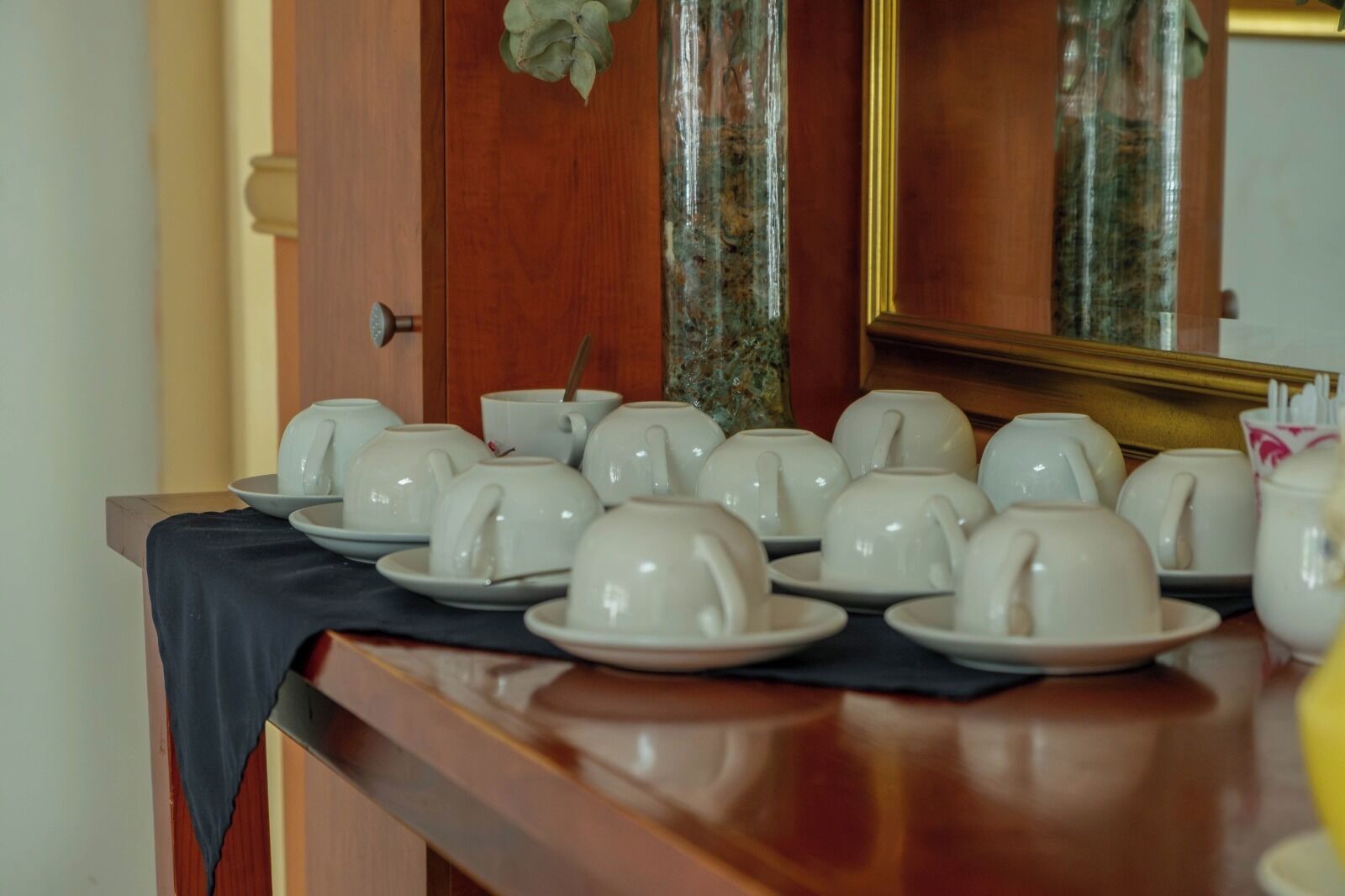 Una mesa dispuesta con teteras y tazas blancas, un mantel negro y un espejo que refleja la habitación.