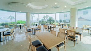 A spacious indoor dining area with multiple tables and chairs, large windows offering a view of palm trees and a body of water. - Seaore Resort (Seogwipo)