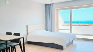 A modern hotel room with a large bed, a dining table with chairs, and a balcony with a view of the ocean. - Seaore Resort (Seogwipo)