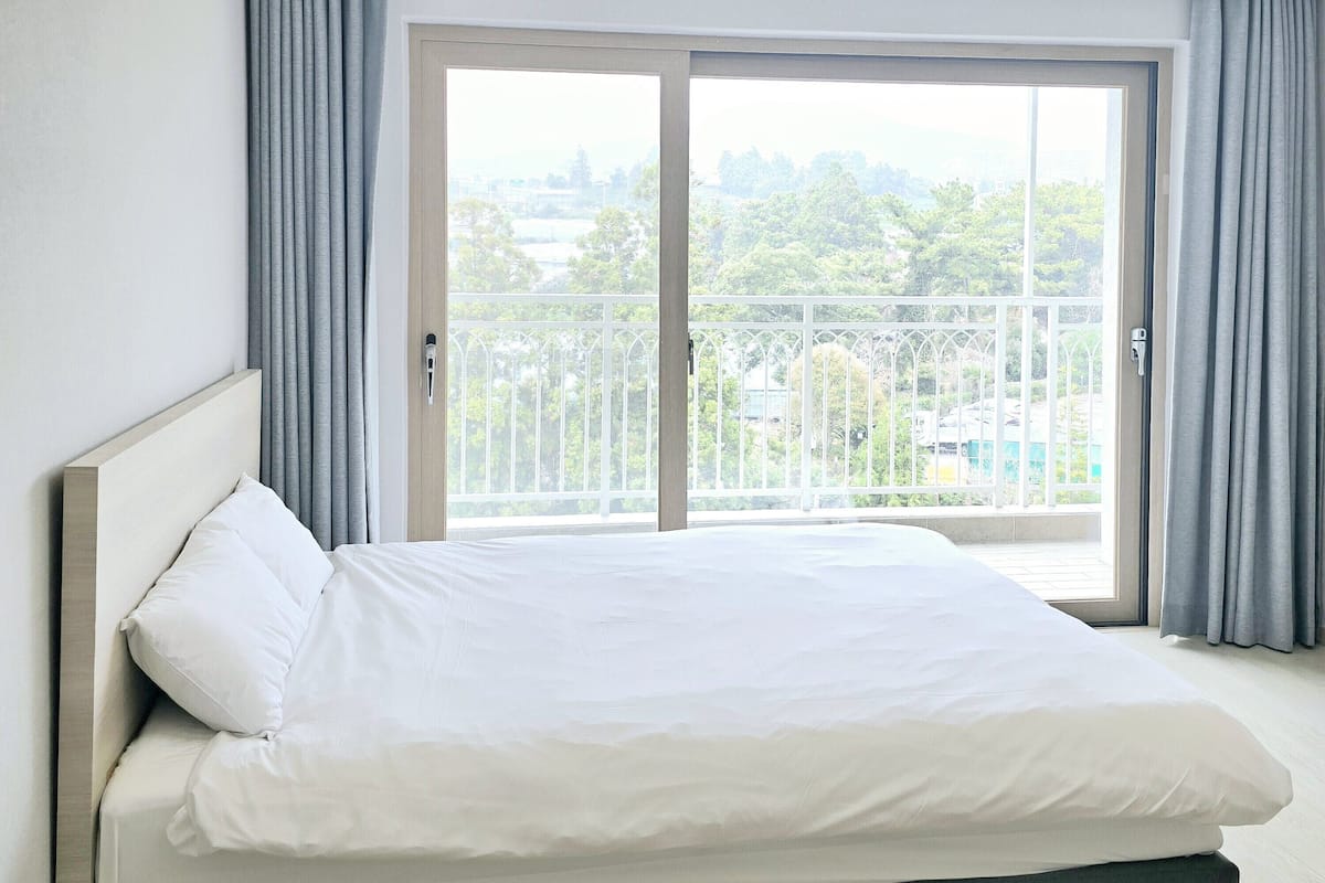 A modern hotel room with a large bed, a balcony with a view of greenery, and a sliding glass door.