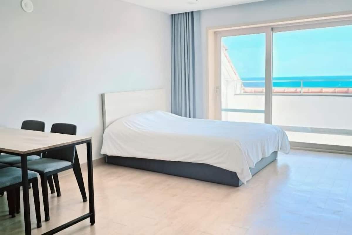 A hotel room with a bed, a table with chairs, and a large window with a view of the sea.