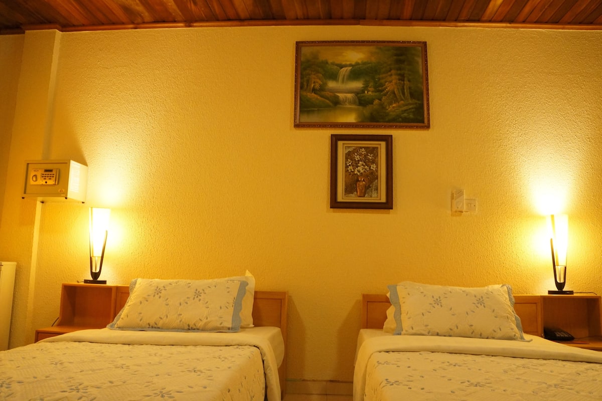 Une chambre d’hôtel avec deux lits, chacun étant doté d’une table de chevet et d’une lampe. Un tableau est accroché au mur, au-dessus des lits.