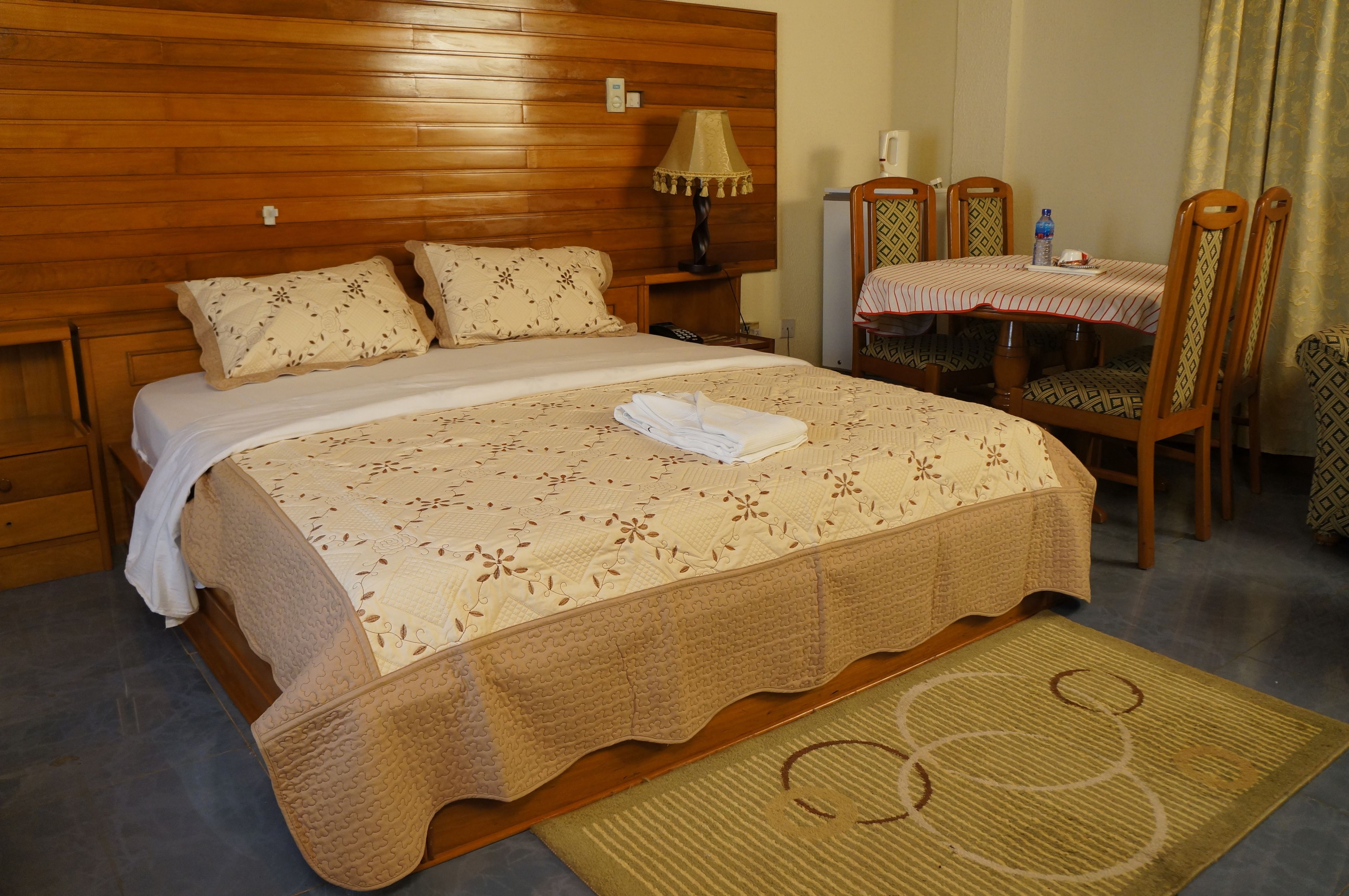 Un lit bien fait, recouvert d’une couvre-lit à motifs floraux, une table de chevet avec une lampe, une table à manger avec des chaises et un tapis tissé sur le sol.