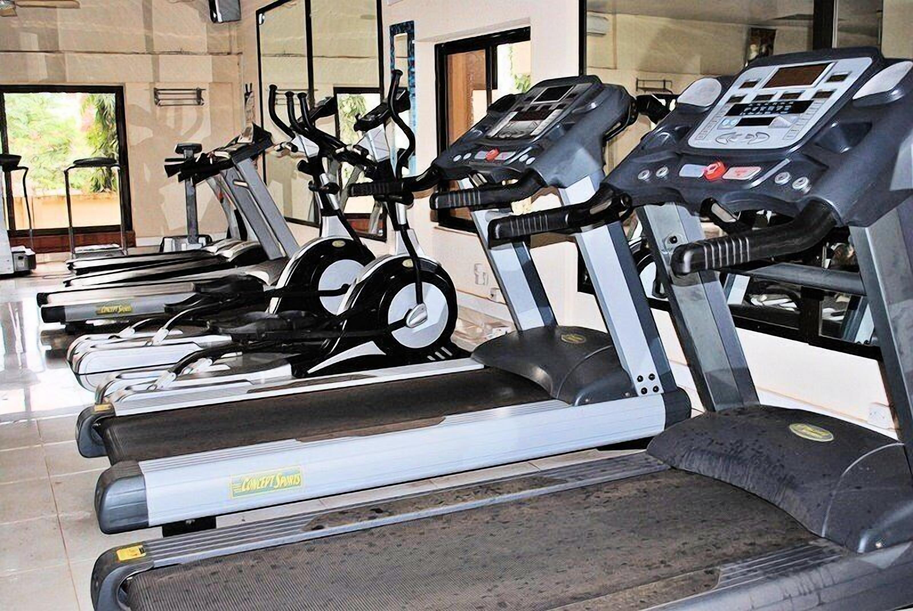A row of stationary exercise bikes in a gym with a mirrored wall and a window in the background.