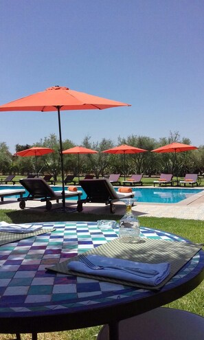 A poolside area with lounge chairs and red umbrellas, a checkered table with a glass and napkin, and a clear blue sky. - Le Diamant De Zaraba (Marrakech)