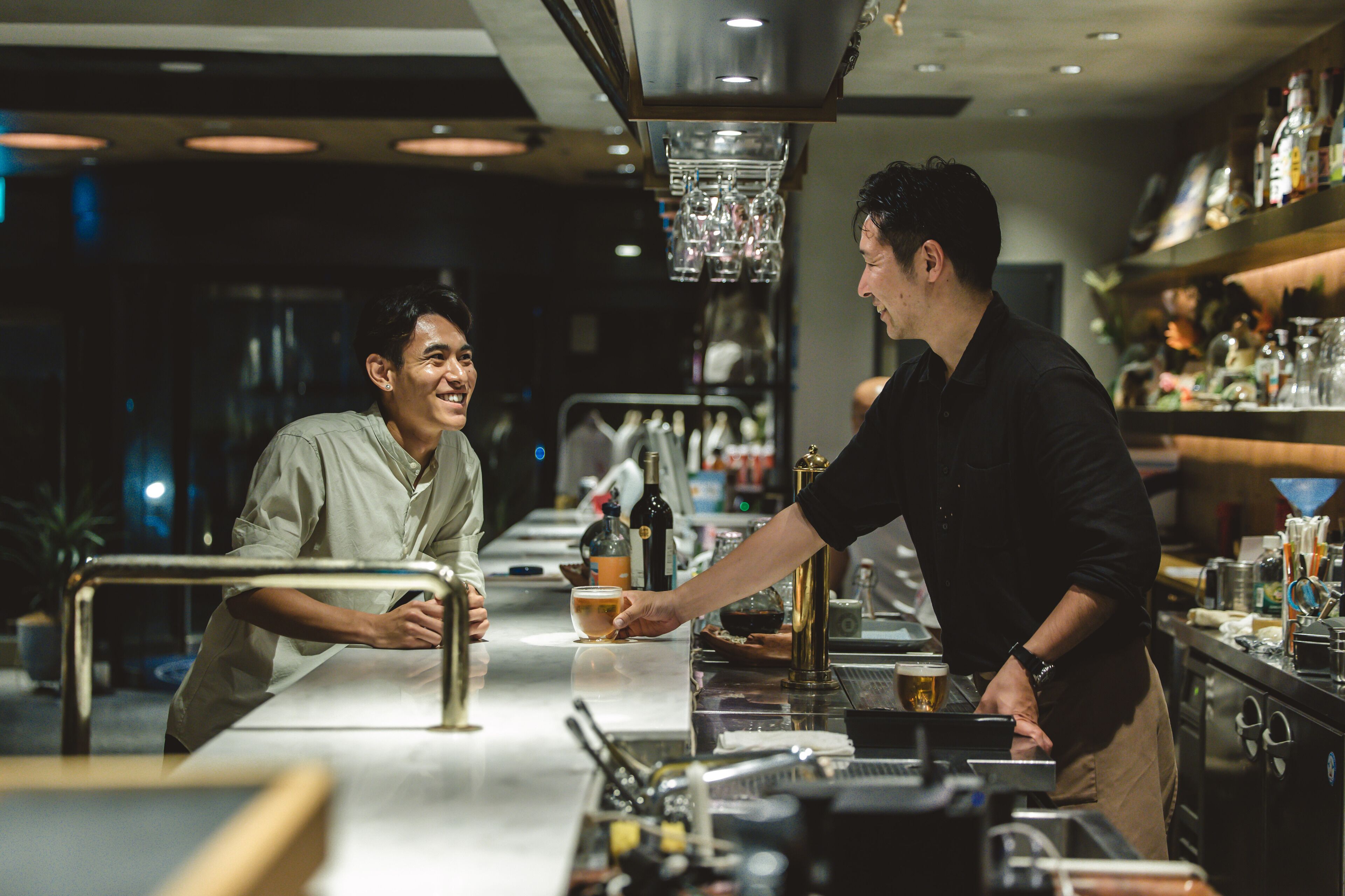 A bar area with a bartender serving a customer. There are bottles and glasses on the counter, and a hanging light fixture above.