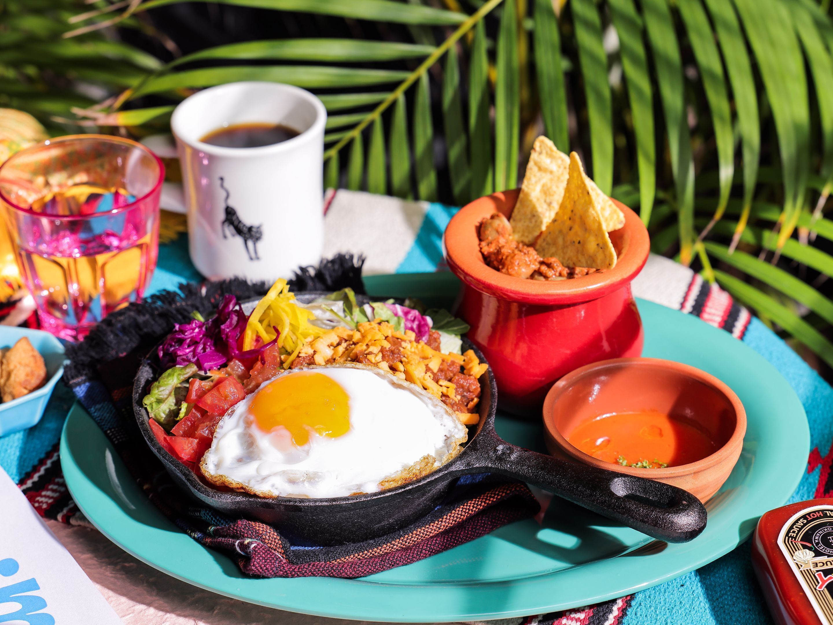A breakfast plate with eggs, vegetables, and chips on a turquoise plate, accompanied by a coffee cup and a bowl of soup.