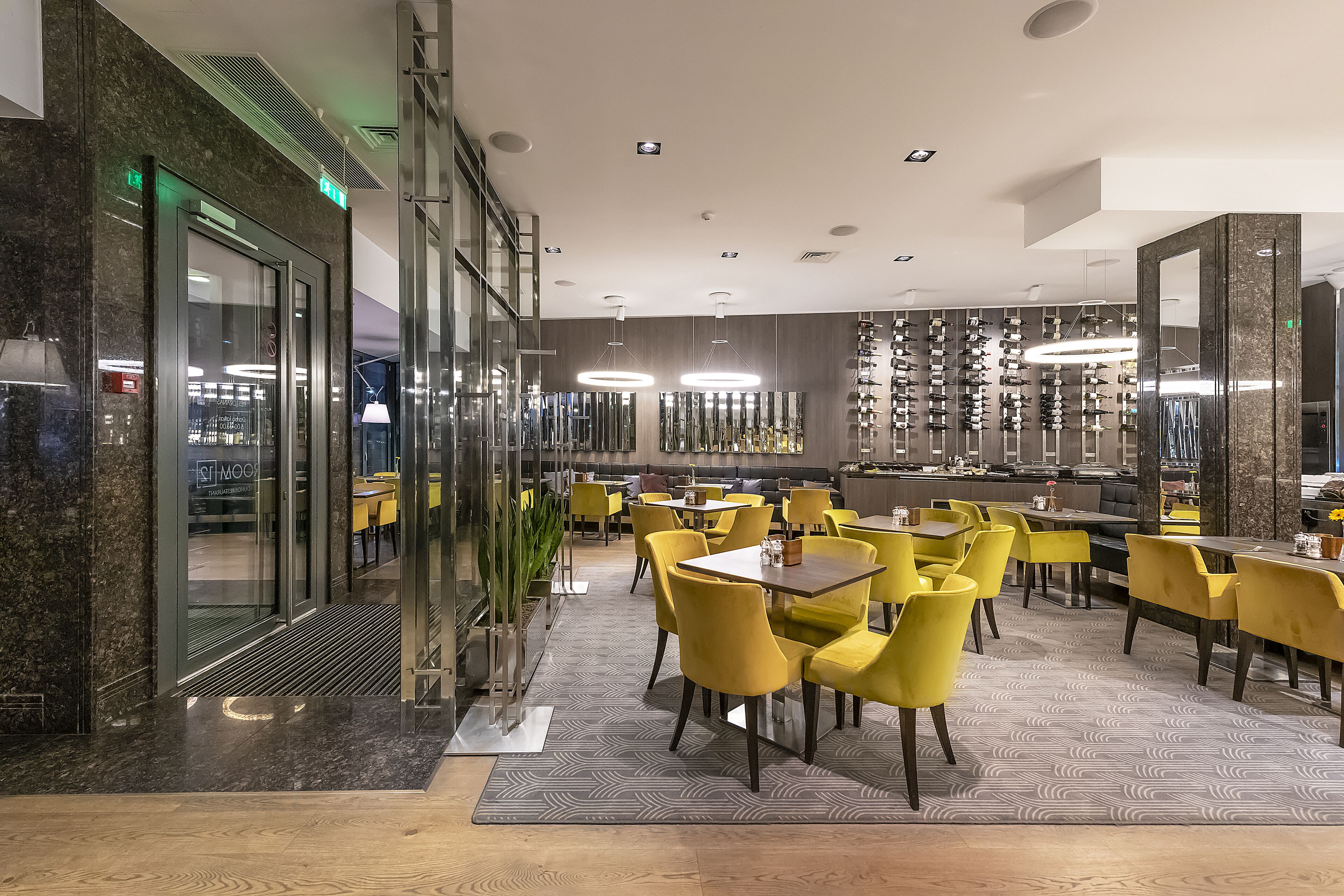 Een modern restaurant met gele stoelen en tafels, een glazen wand en een wand vol hangende glazen.