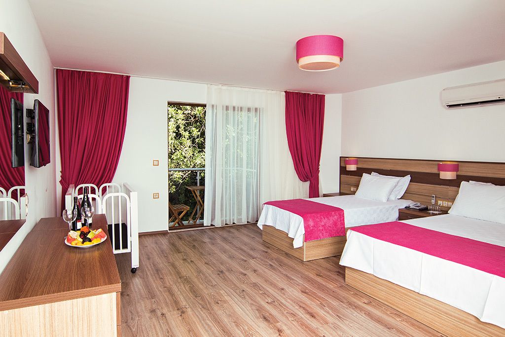 A hotel room with two beds, a wooden desk, a dining table with fruit, and a balcony with red curtains.