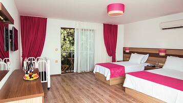 A hotel room with two beds, a wooden desk, a dining table with fruit, and a balcony with red curtains.