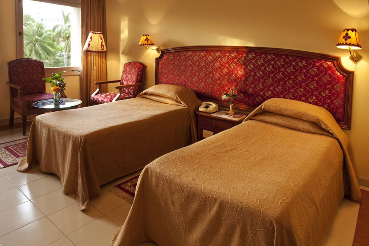 A hotel room with two single beds, a sitting area with a chair and a small table, and a window with curtains.