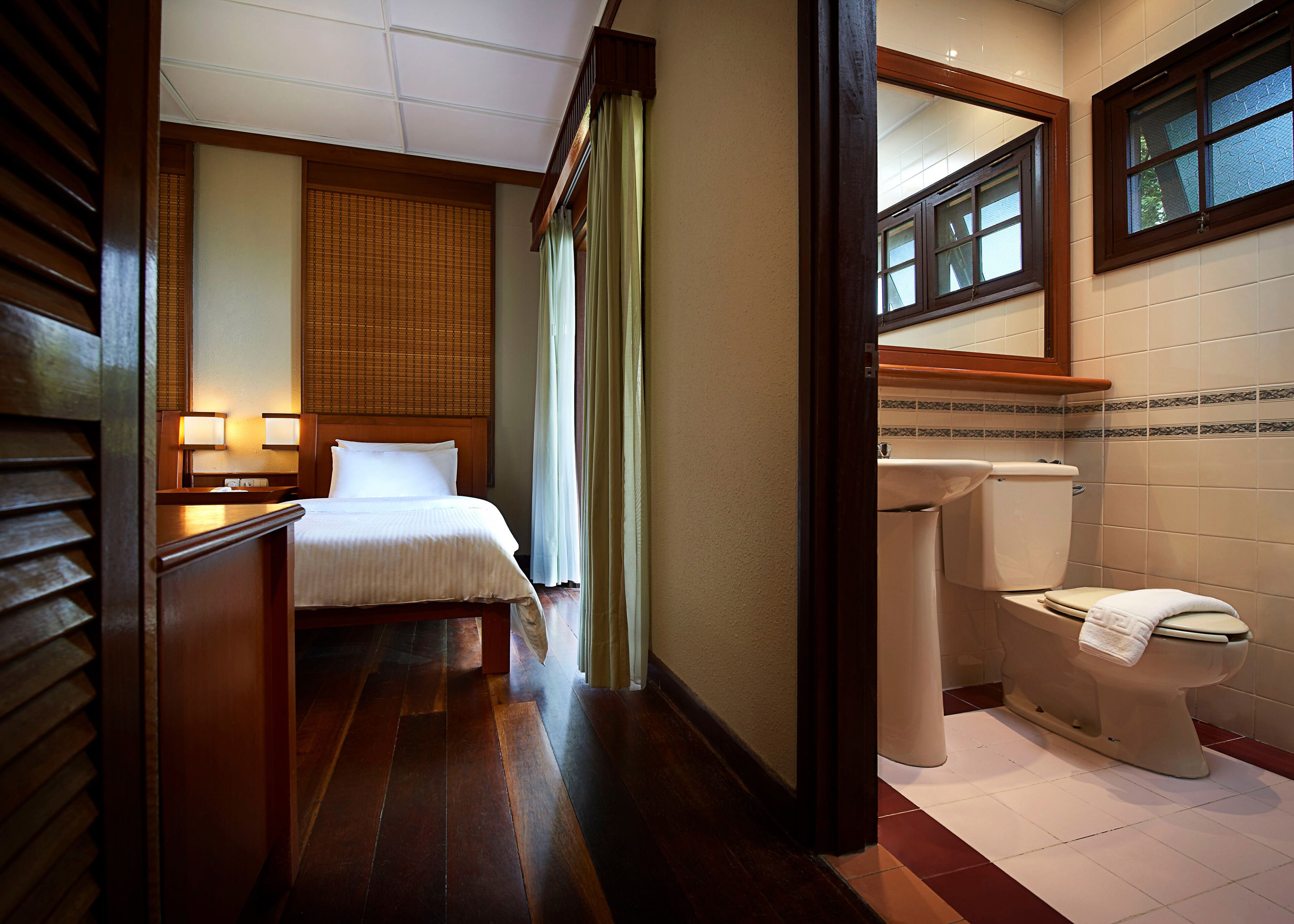 A hotel room with a bed, bedside table, lamp, window, and a bathroom with a toilet and sink.