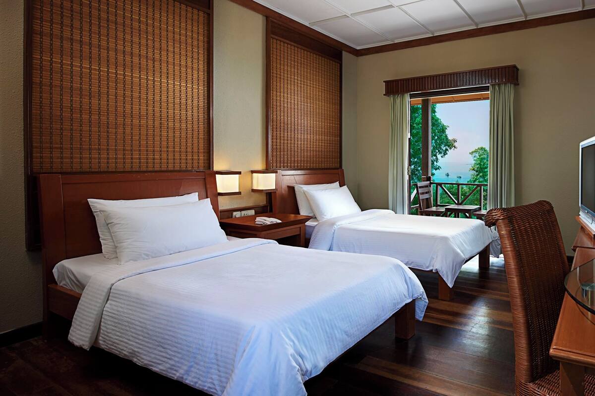 A hotel room with two beds, a TV, and a balcony with a view of the sea.