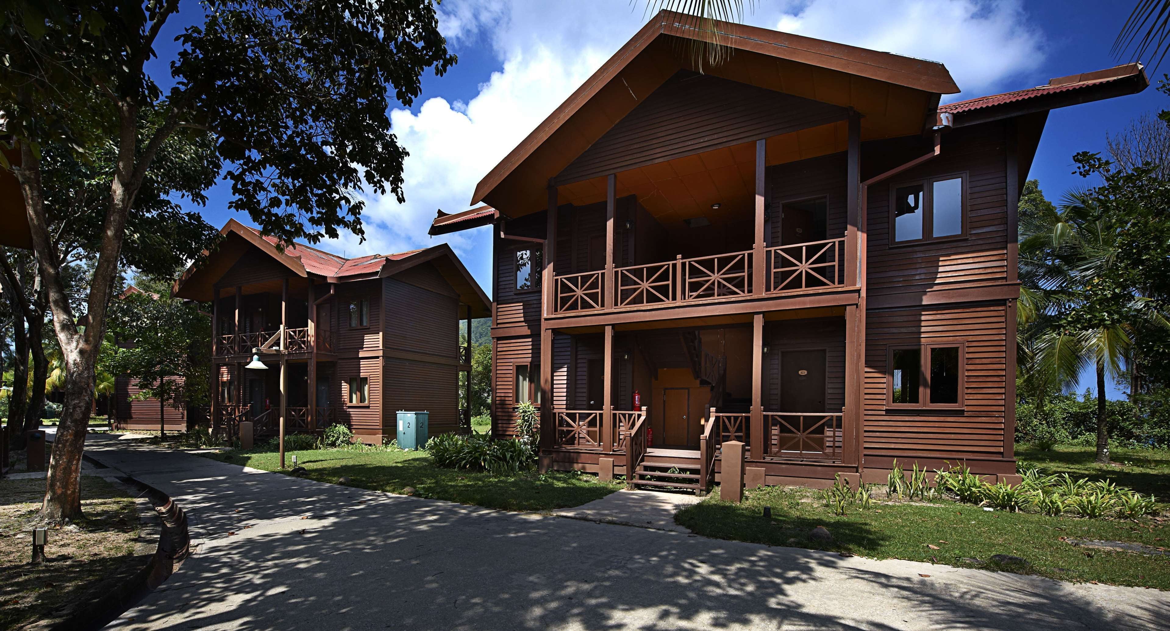 A two-story wooden cabin with a balcony, surrounded by greenery and a clear sky.
