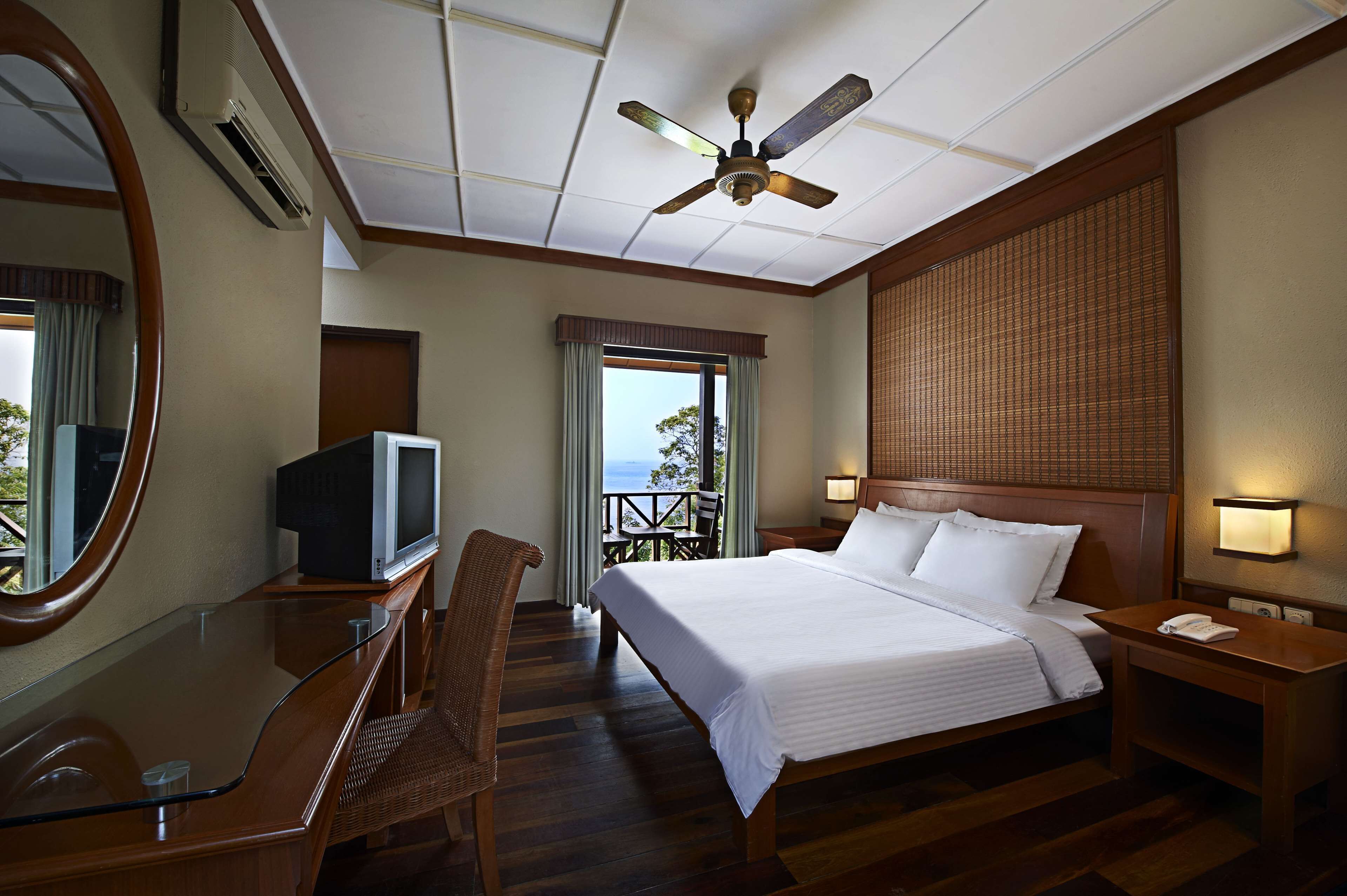 Quarto de hotel com cama, televisão, escrivaninha, cadeira, varanda com vista e ventoinha no teto.