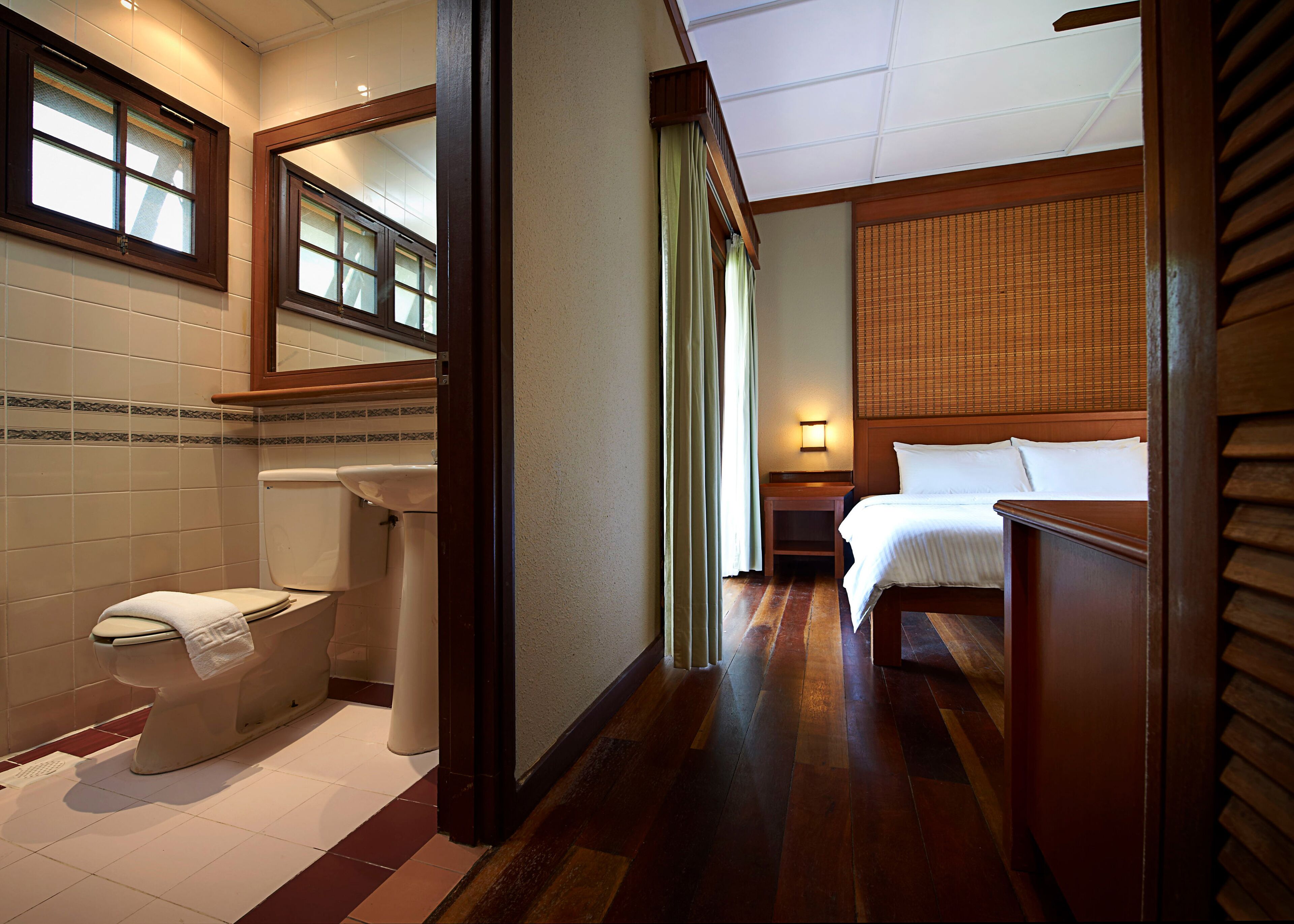 A hotel room with a wooden floor, a bed, a nightstand, a bathroom with a toilet, and a window with blinds.