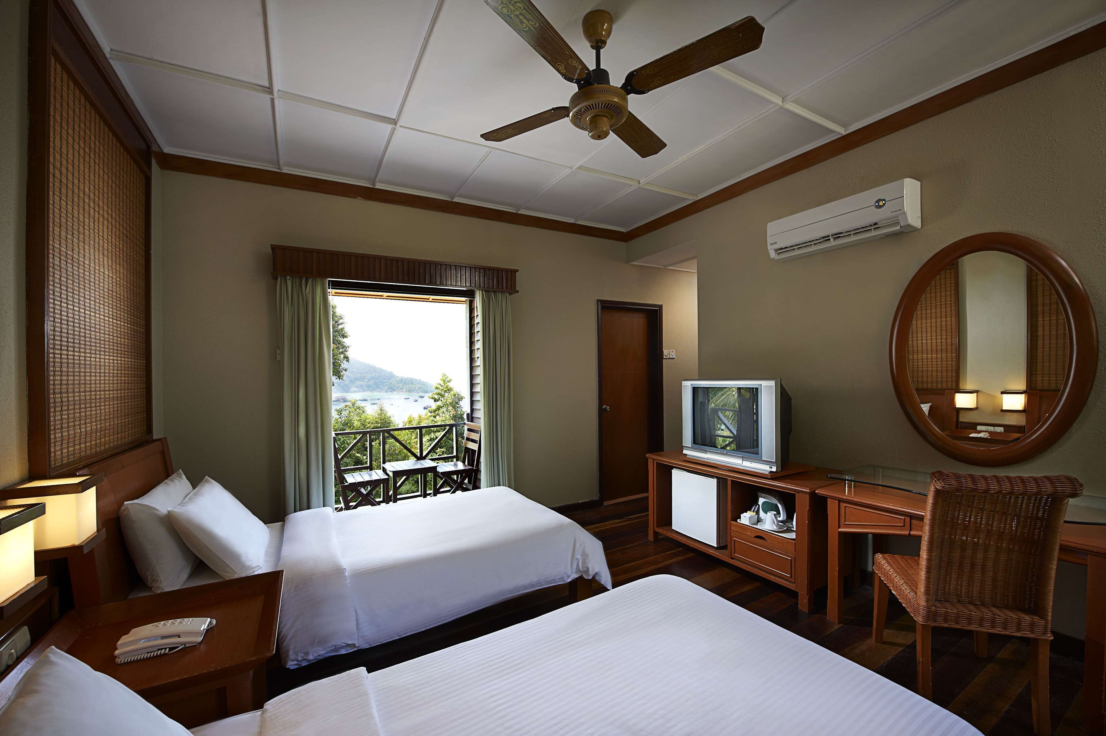 Habitación de hotel con dos camas, televisión, escritorio, silla, ventilador de techo y vistas al exterior.