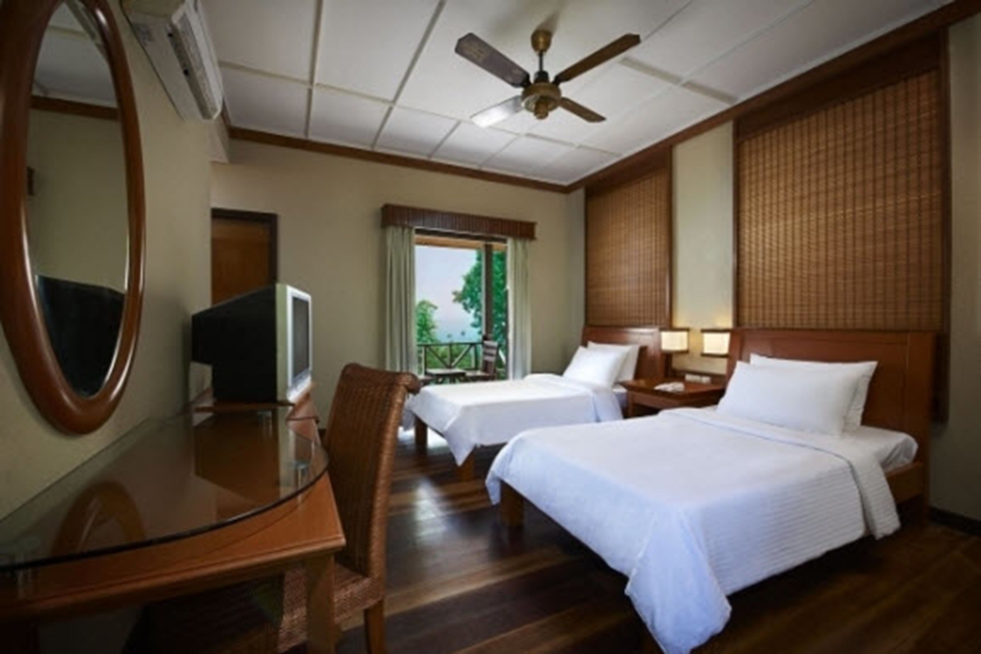 A hotel room with two beds, a desk, a chair, a television, a ceiling fan, and a window with a view of greenery.