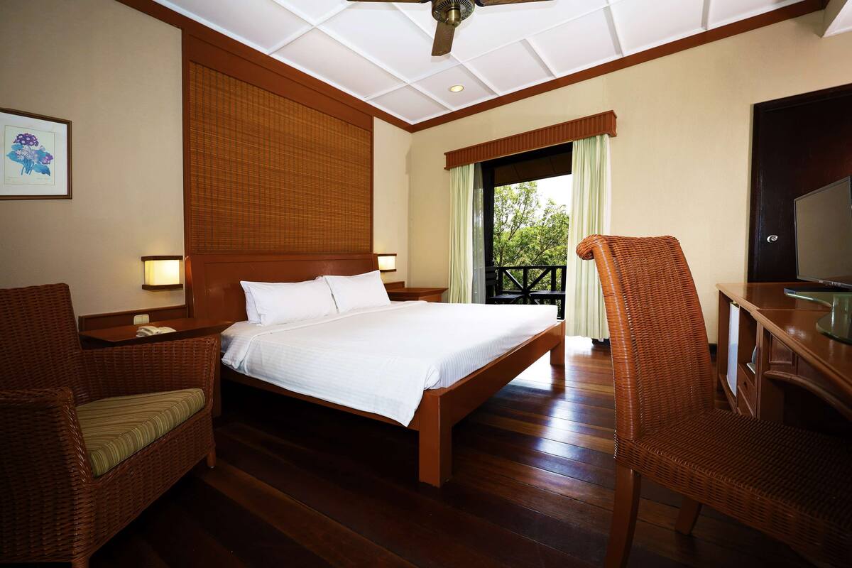 A hotel room with a wooden bed, two wicker chairs, a desk with a television, and a large window with a view of greenery.