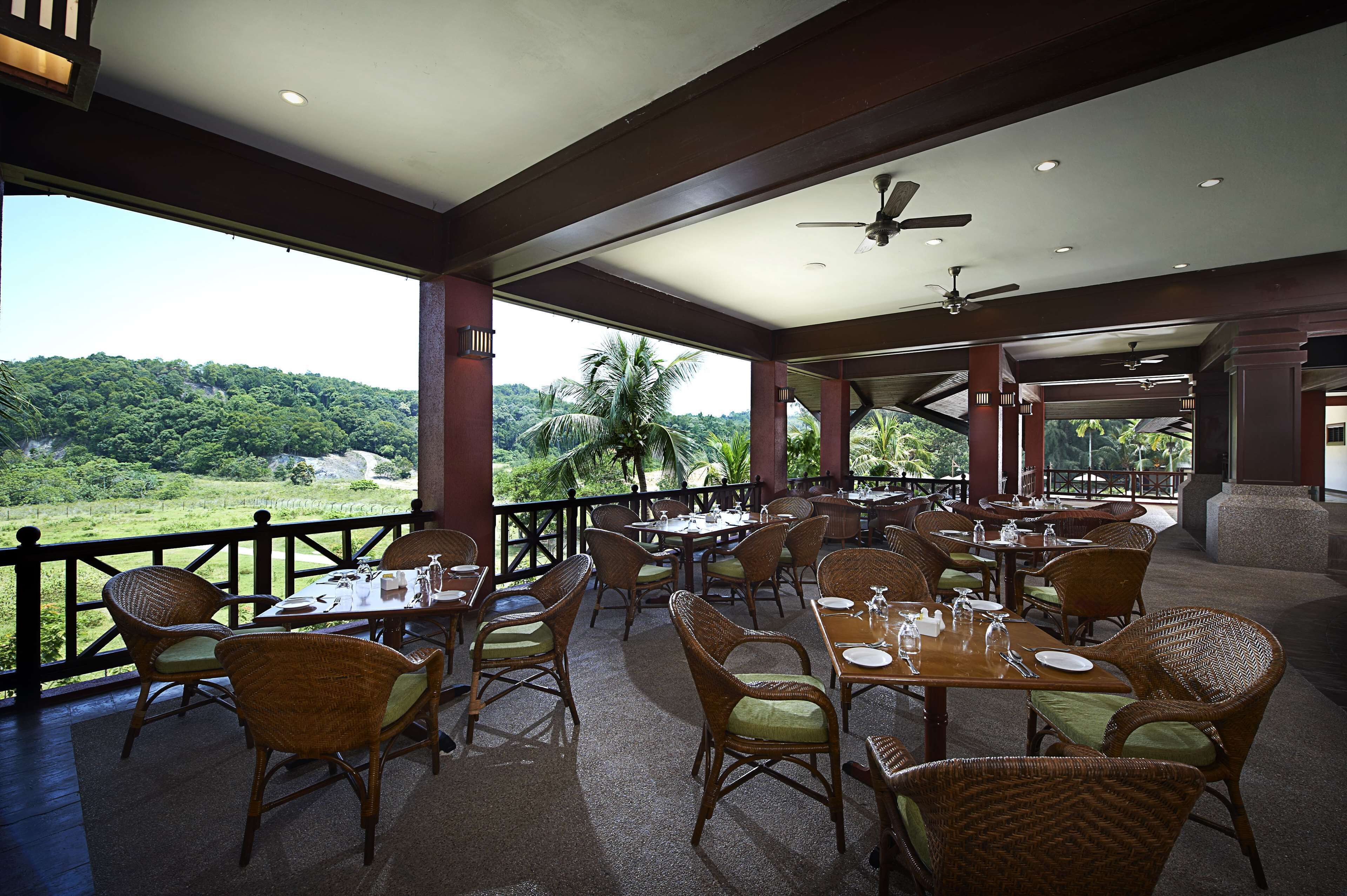 A restaurant with wicker chairs and tables, a ceiling fan, and a view of a lush green landscape.