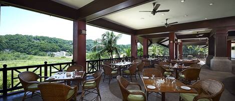 A restaurant with wicker chairs and tables, a ceiling fan, and a view of a lush green landscape.