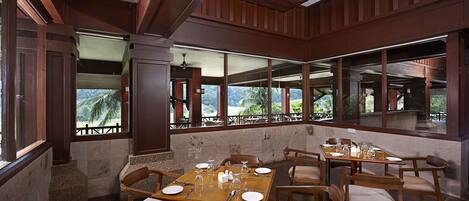 A dining area with a large table set for six, surrounded by wooden chairs with cushioned seats. The room features large windows offering a view of a mountainous landscape.