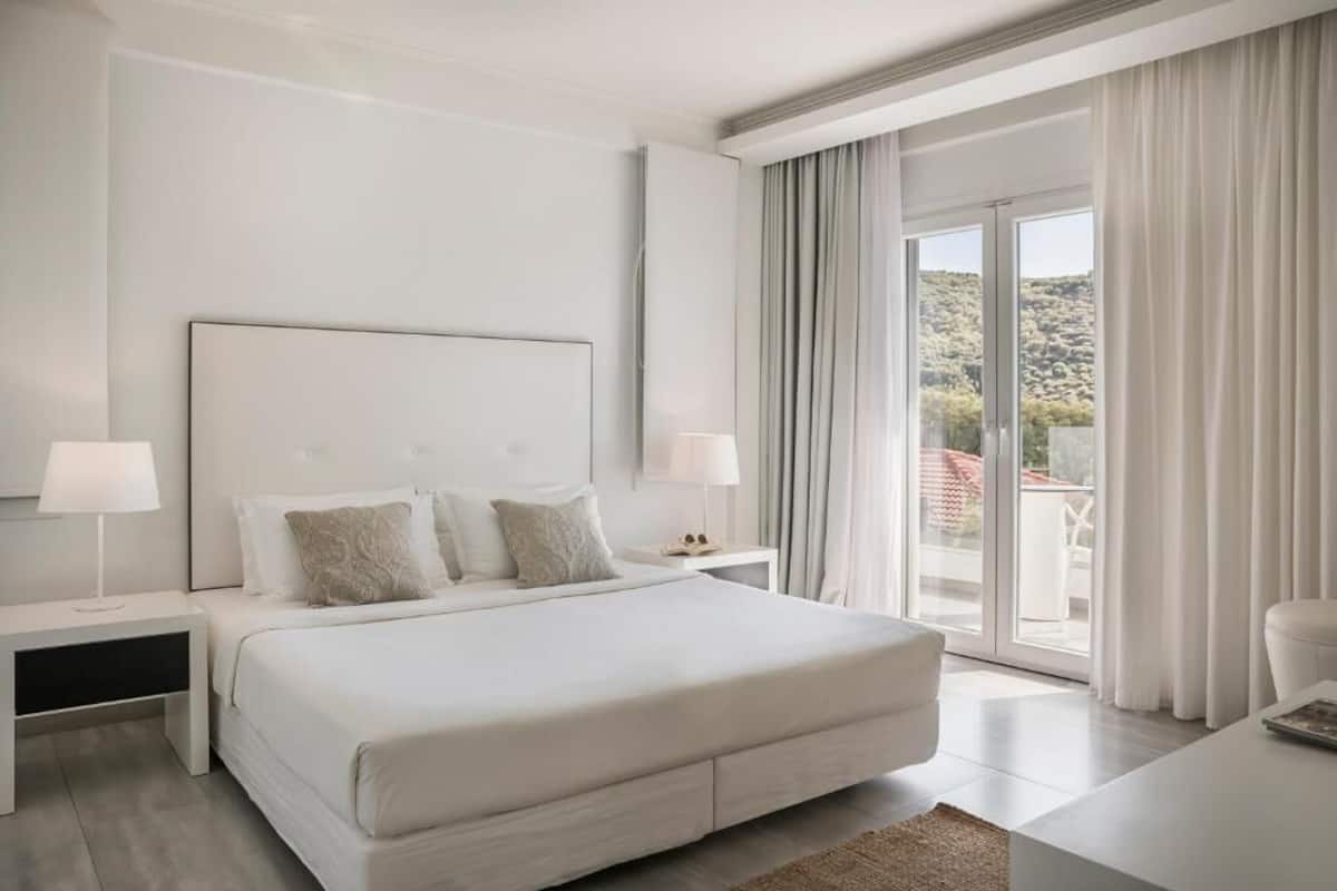 A modern hotel room with a large bed, two bedside tables, a nightstand, and a view of the outdoors through a sliding glass door.