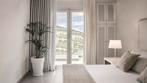Une chambre d’hôtel moderne avec un grand lit, une table de chevet avec une lampe, une plante en pot et une vue sur un paysage montagneux, visible à travers une porte coulissante.