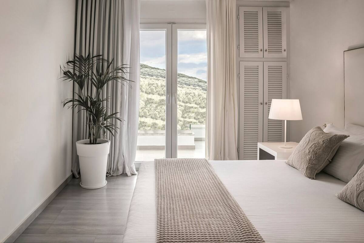 A modern hotel room with a large bed, a bedside table with a lamp, a potted plant, and a view of a mountainous landscape through a sliding door.