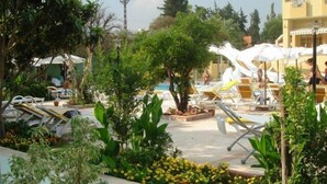Un espace aménagé au bord de la piscine, avec des chaises longues et des parasols, entouré de verdure et avec un bâtiment en arrière-plan.