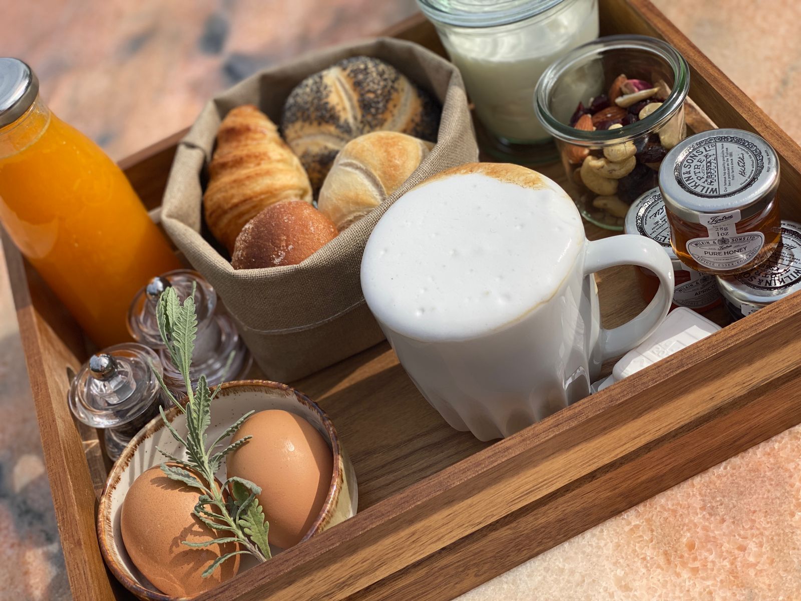 Een houten dienblad met diverse ontbijtartikelen, waaronder croissants, een glas sinaasappelsap, een mok koffie, een pot honing en een schaal met noten.
