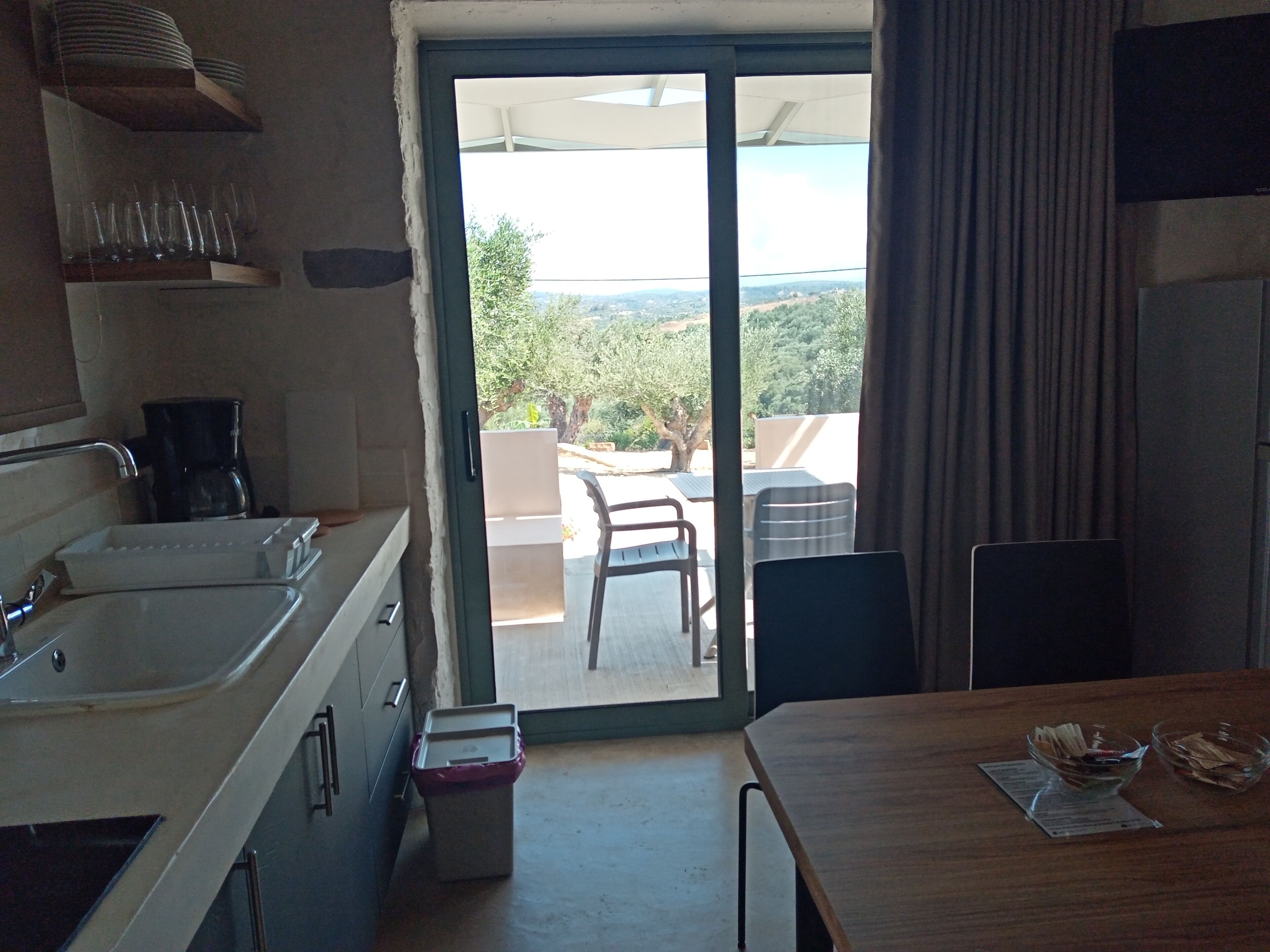 A kitchen with a large sink, a coffee maker, and a trash bin. There is a dining table with chairs and a view of the outdoors through a sliding door.