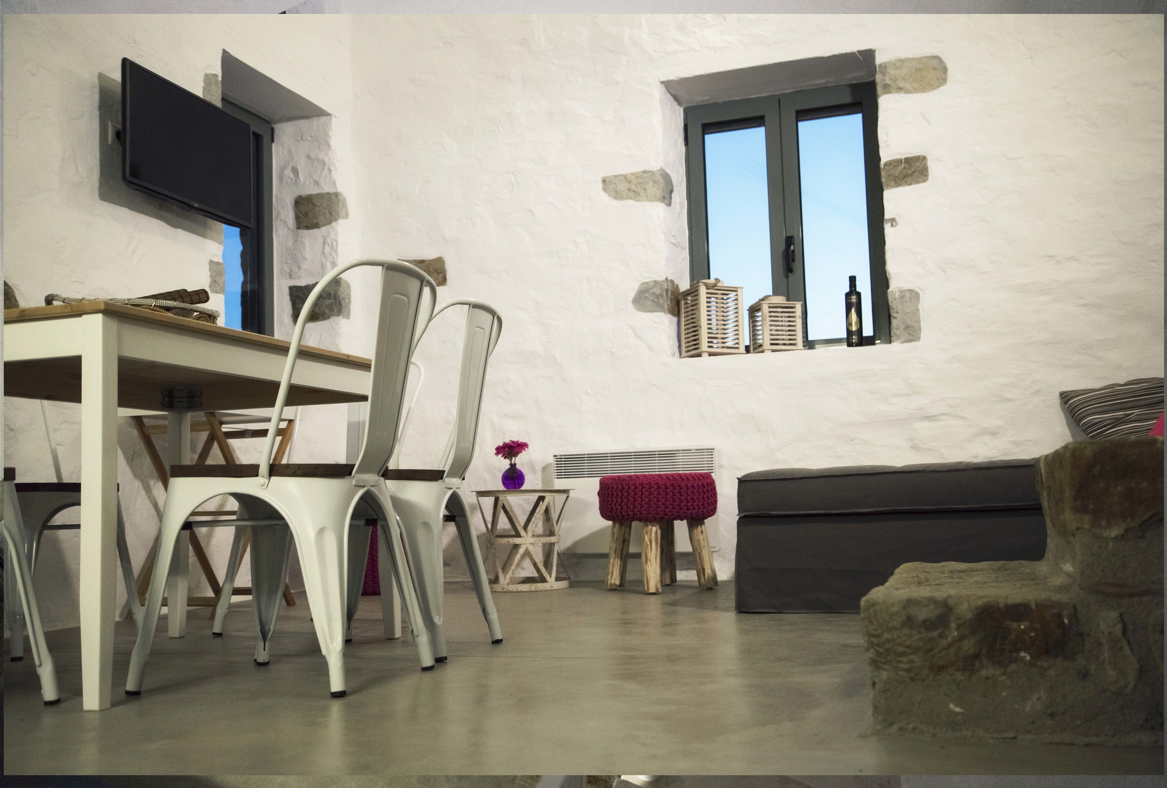 Ein Zimmer mit Steinwand, einem an der Wand über einer Tür befestigten Fernseher, einem Fenster mit Körben, einem Esstisch mit Stühlen und einem Sofa.